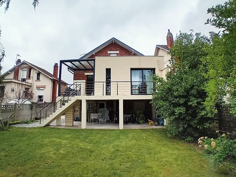 Maison au pignon en brique prolongée par un volume enduit beige à toit plat, grande terrasse sur poteaux à garde-corps anthracite, escalier droit en béton vers une pelouse, pergola métallique à gauche.