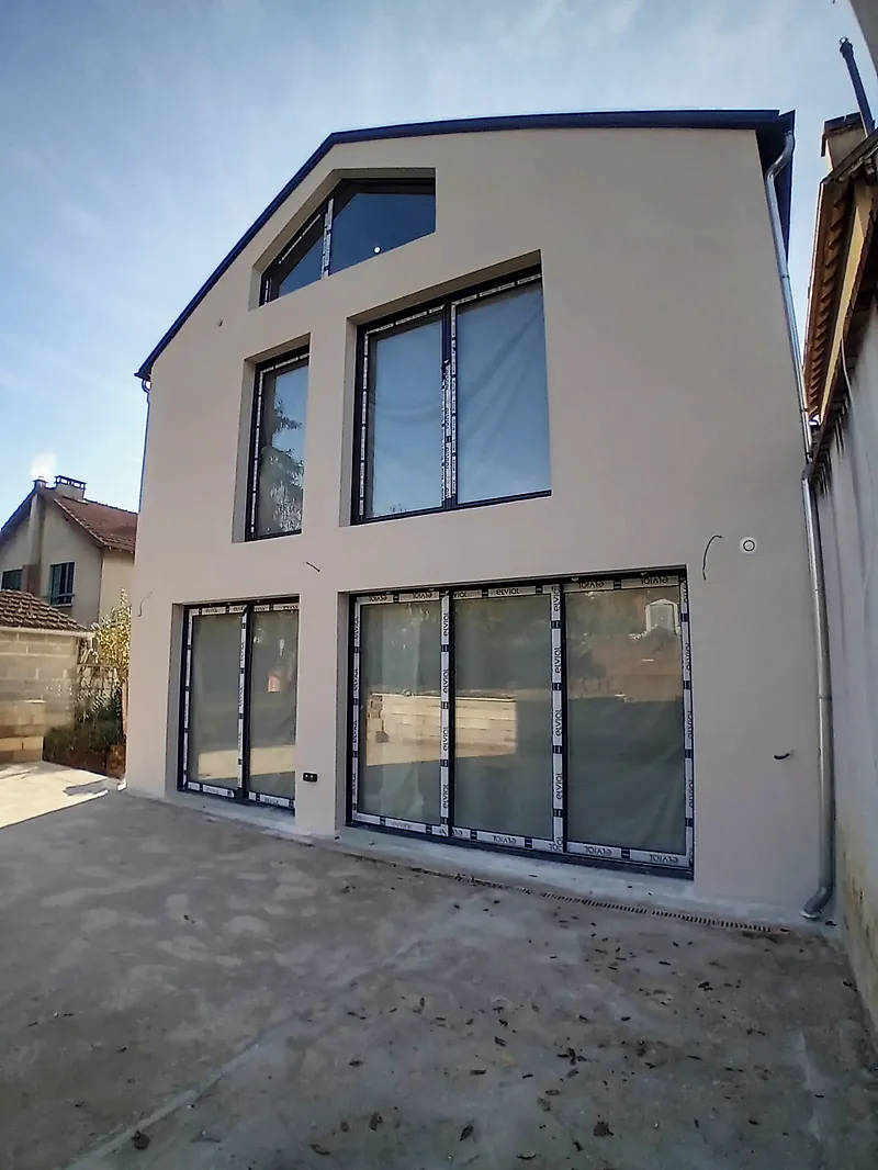 Façade-pignon contemporaine enduite gris clair, rives sombres, grandes baies vitrées noires au rez-de-chaussée, deux fenêtres à l’étage et une baie trapézoïdale sous le toit.