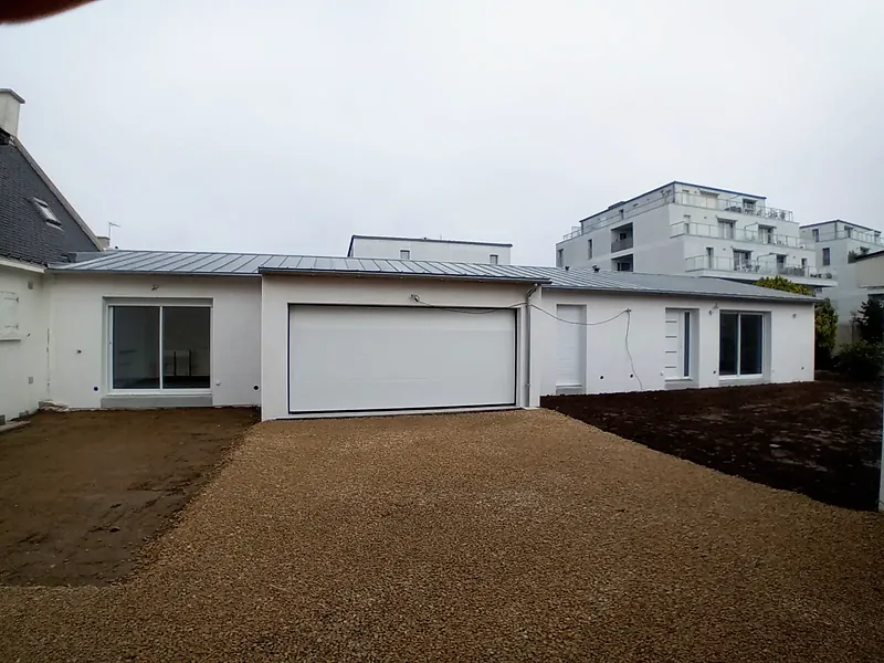 Extension de plain-pied enduite de blanc avec grande porte de garage sectionnelle blanche, baies coulissantes et couverture métallique grise à joints debout, cour gravillonnée au premier plan, résidences récentes en arrière-plan.