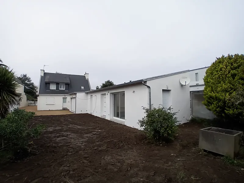 Long volume de plain-pied enduit de blanc, baie coulissante et fenêtres, maison à toit d’ardoise et lucarnes au fond, jardin en terre fraîchement nivelée.
