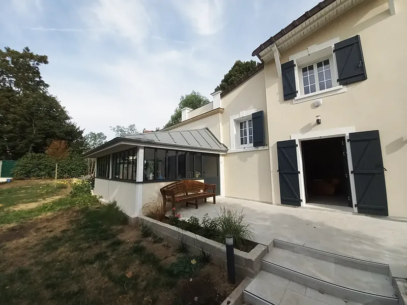 Terrasse en pierre claire devant une double porte encadrée de volets gris-bleu, extension vitrée et banc en bois à gauche, marches et borne lumineuse au premier plan.