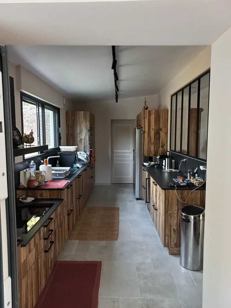 Cuisine en longueur aux façades en bois brut et plan de travail noir, verrière à profils noirs à droite, rail d’éclairage au plafond, carrelage clair.