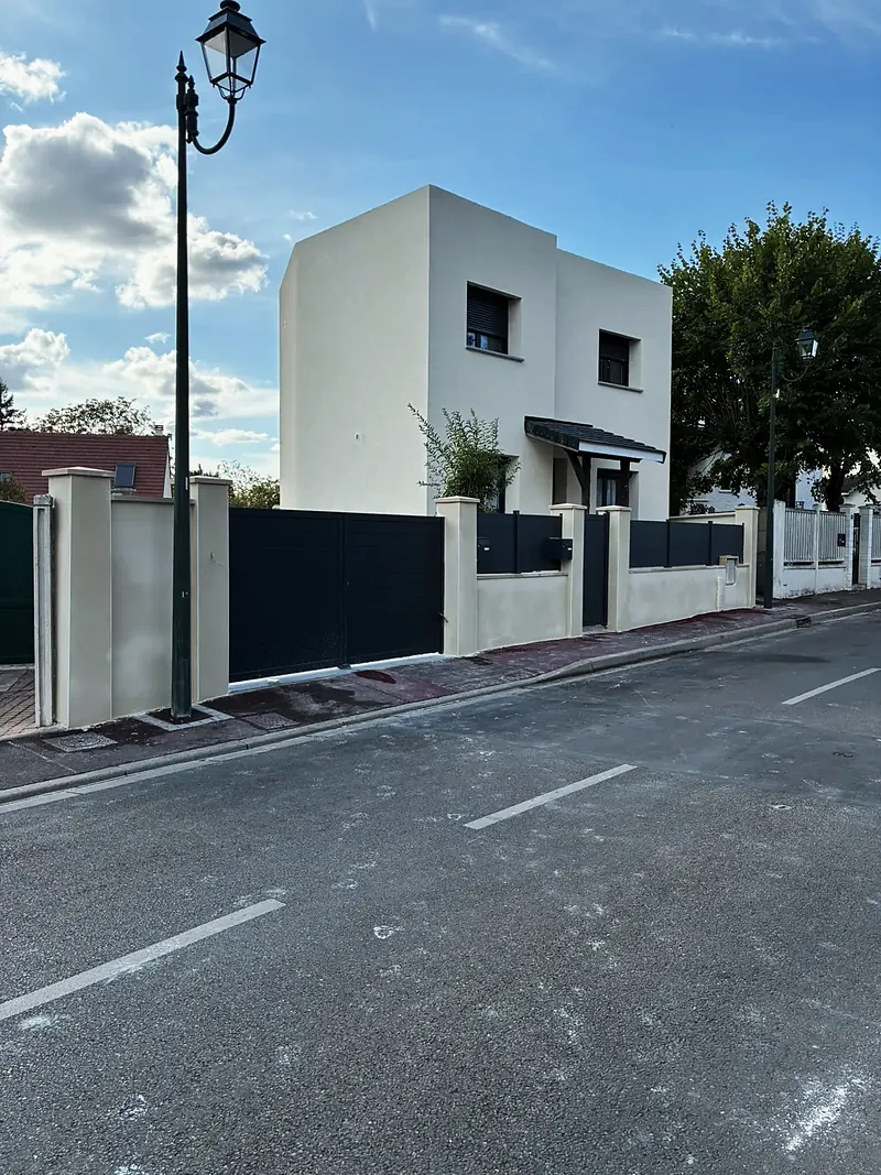 Maison cubique blanche à un étage, deux volumes décalés, menuiseries anthracite, clôture à piliers enduits et portail anthracite, ciel bleu.