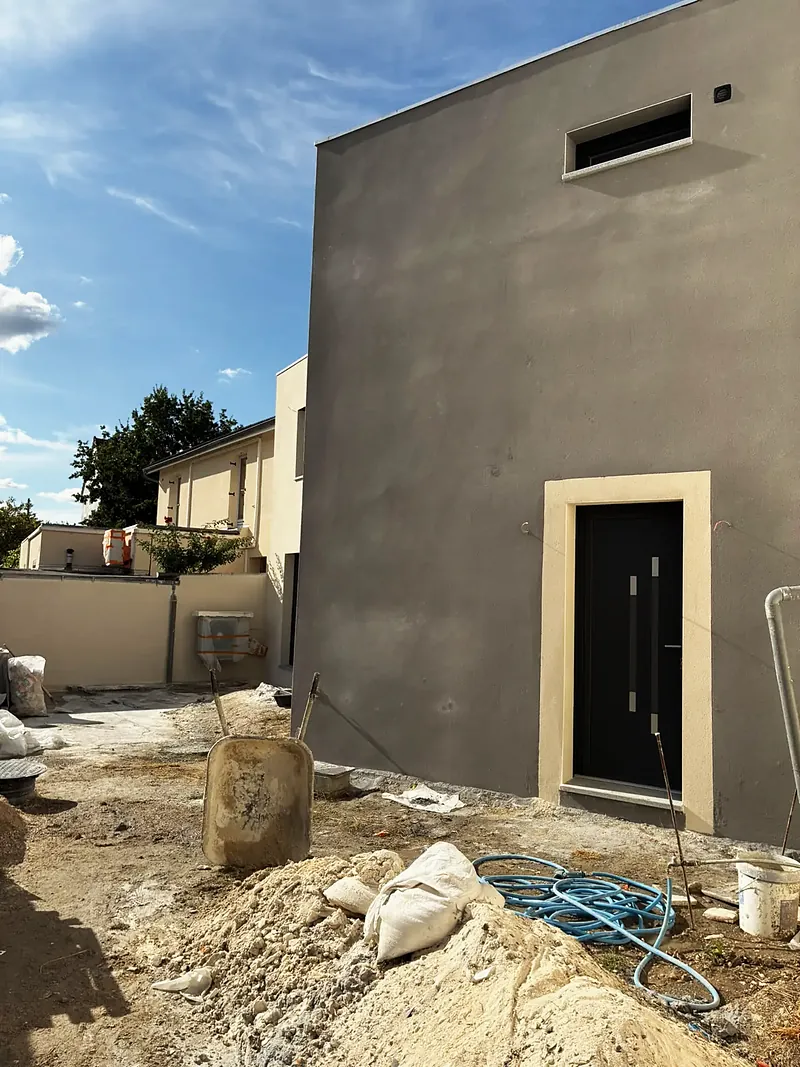 Façade gris foncé avec porte noire encadrée de clair, petite fenêtre haute, jardin en travaux.