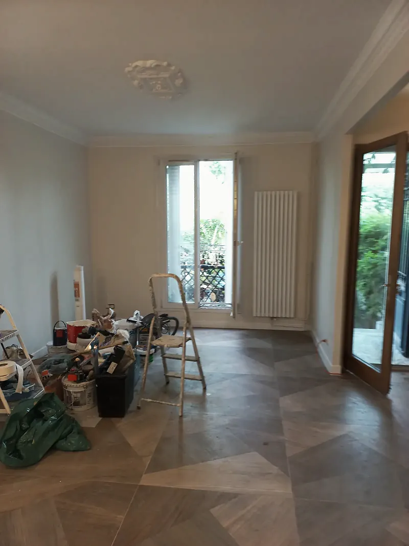 Pièce aux murs clairs avec corniche moulurée et rosace au plafond, radiateur vertical blanc, porte-fenêtre ouverte sur un jardin, pots de peinture et escabeau au sol.