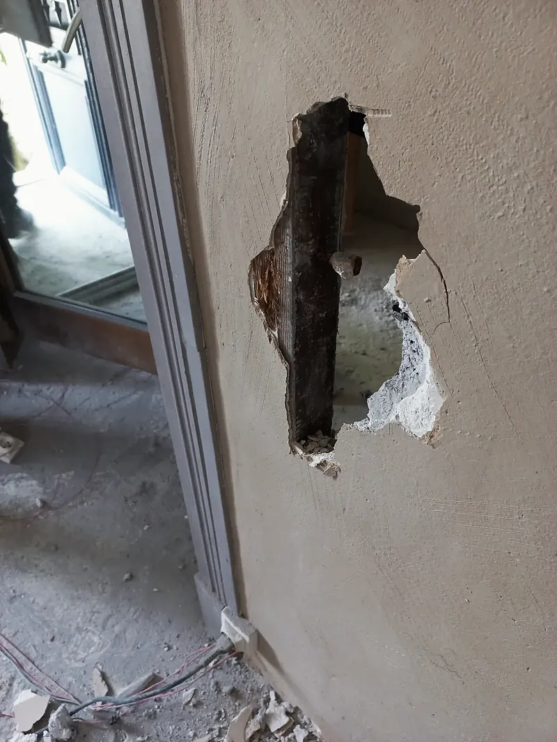 Mur enduit percé d’un trou laissant voir un profilé métallique rouillé, à côté d’un encadrement de porte gris.