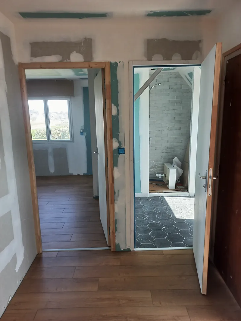 Dégagement au sol en lames ton bois, deux portes ouvertes : à gauche une chambre avec fenêtre, à droite une salle d’eau au sol en carreaux hexagonaux gris foncé.