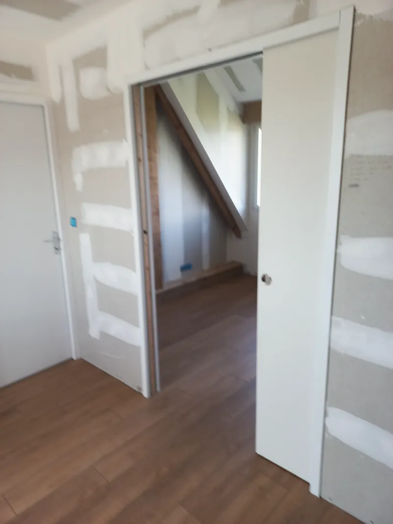 Porte coulissante blanche ouverte dans une cloison en plaques jointoyées, chambre sous rampant au fond, sol en lames ton bois.