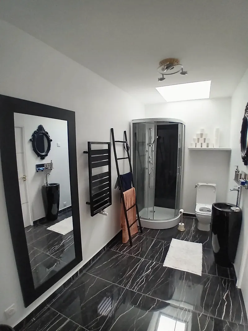Salle d’eau blanche avec puits de lumière au plafond, carrelage noir veiné, cabine de douche et sèche-serviettes noir.